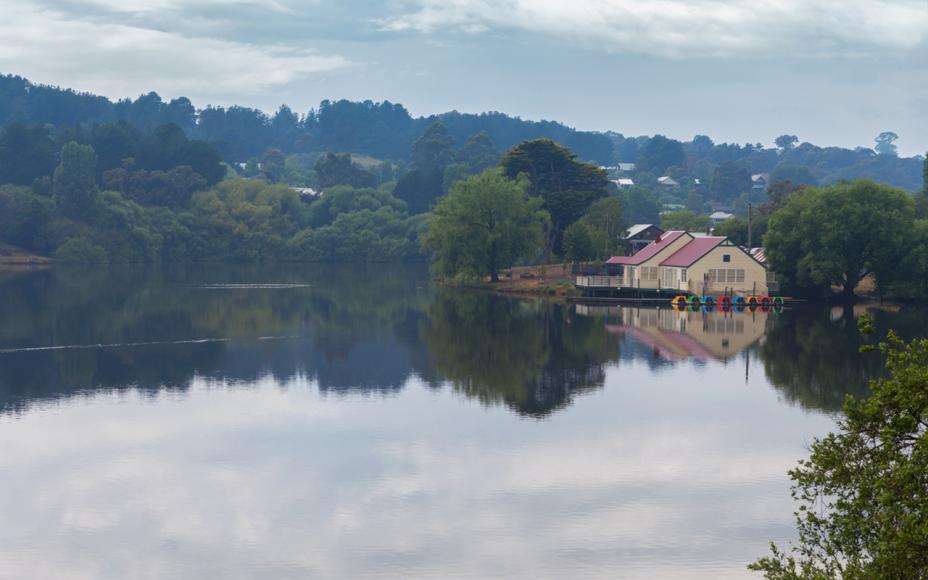 Daylesford lake, Victoria. Peaceful lakeside scene with a boathouse reflected in still water, surrounded by lush green trees and hills under a cloudy sky, conveying tranquility.